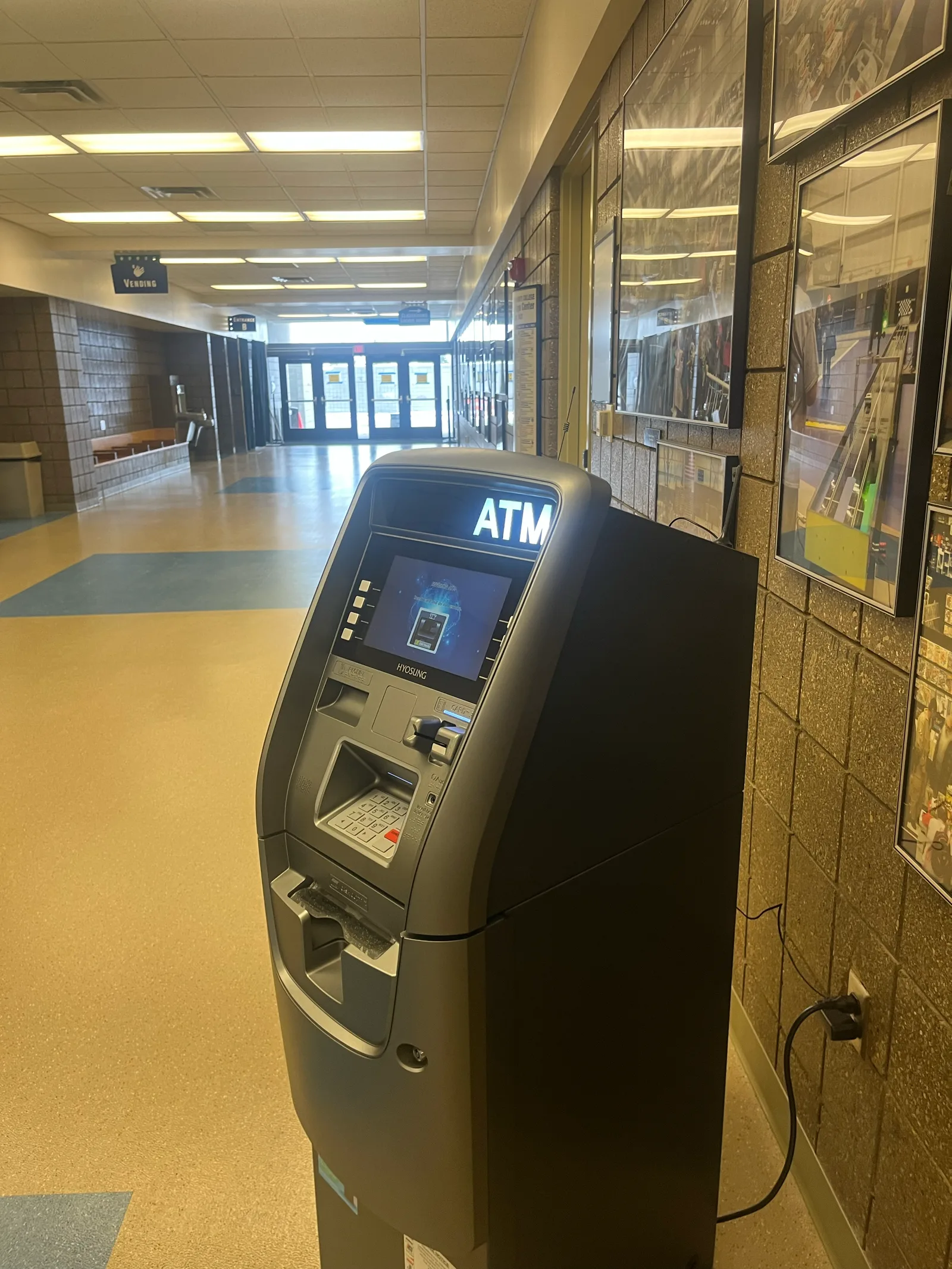 An ATM in a public building corridor with display cases along the wall