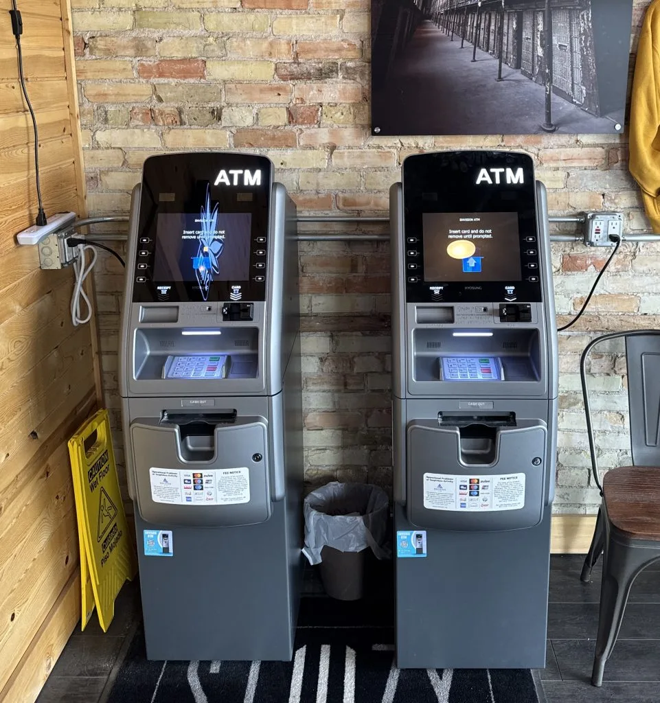 Two Envision ATM machines installed side by side against an exposed brick wall in a bar